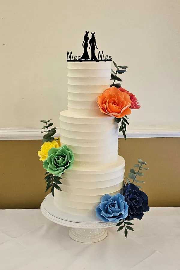 3 tier wedding cake. Buttercream scalloped edging sides, with handmade sugar flowers in rainbow colours, and a Mrs & Mrs acrylic cake topper.